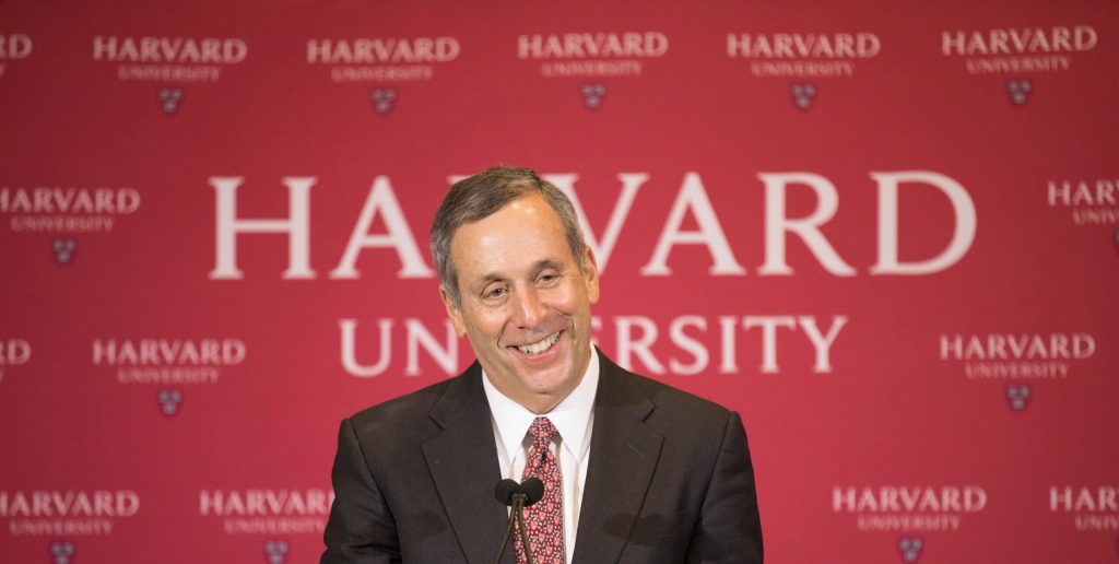 Man in a suit in front of a Harvard University banner.