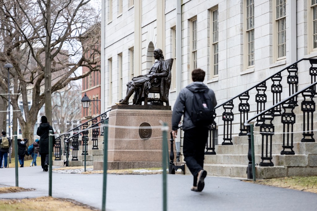 Young man walks in front of the John Harvard statue.