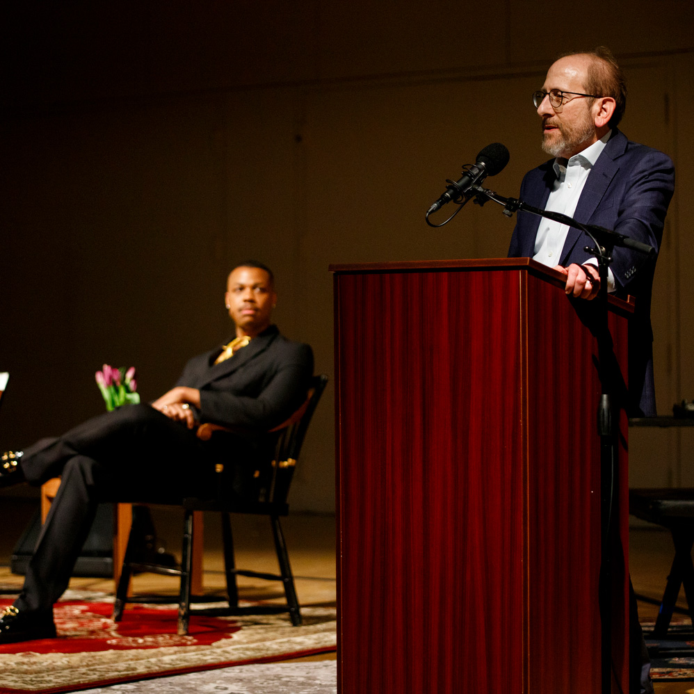 Alan Garber speaks at a podium with Davone Tines in the background looking on.