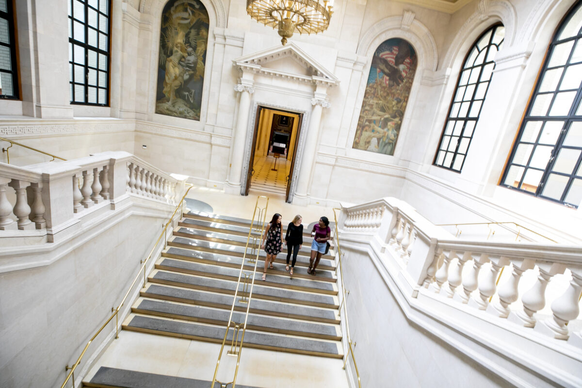 Three young women walking up the stairs, indoors, of a spacious white marble building.