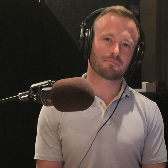 A man wearing headphones sits by a large microphone while recording a podcast
