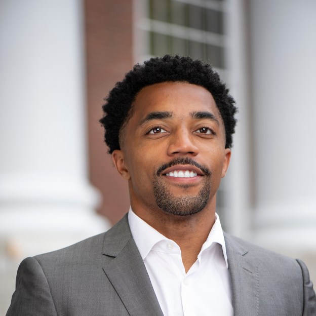 A man wearing a suit stands outside a large brick building with white columns