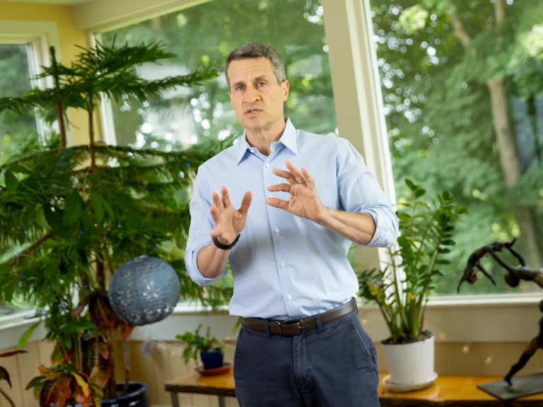 A man talks while standing in a sunny room with indoor plants behind him