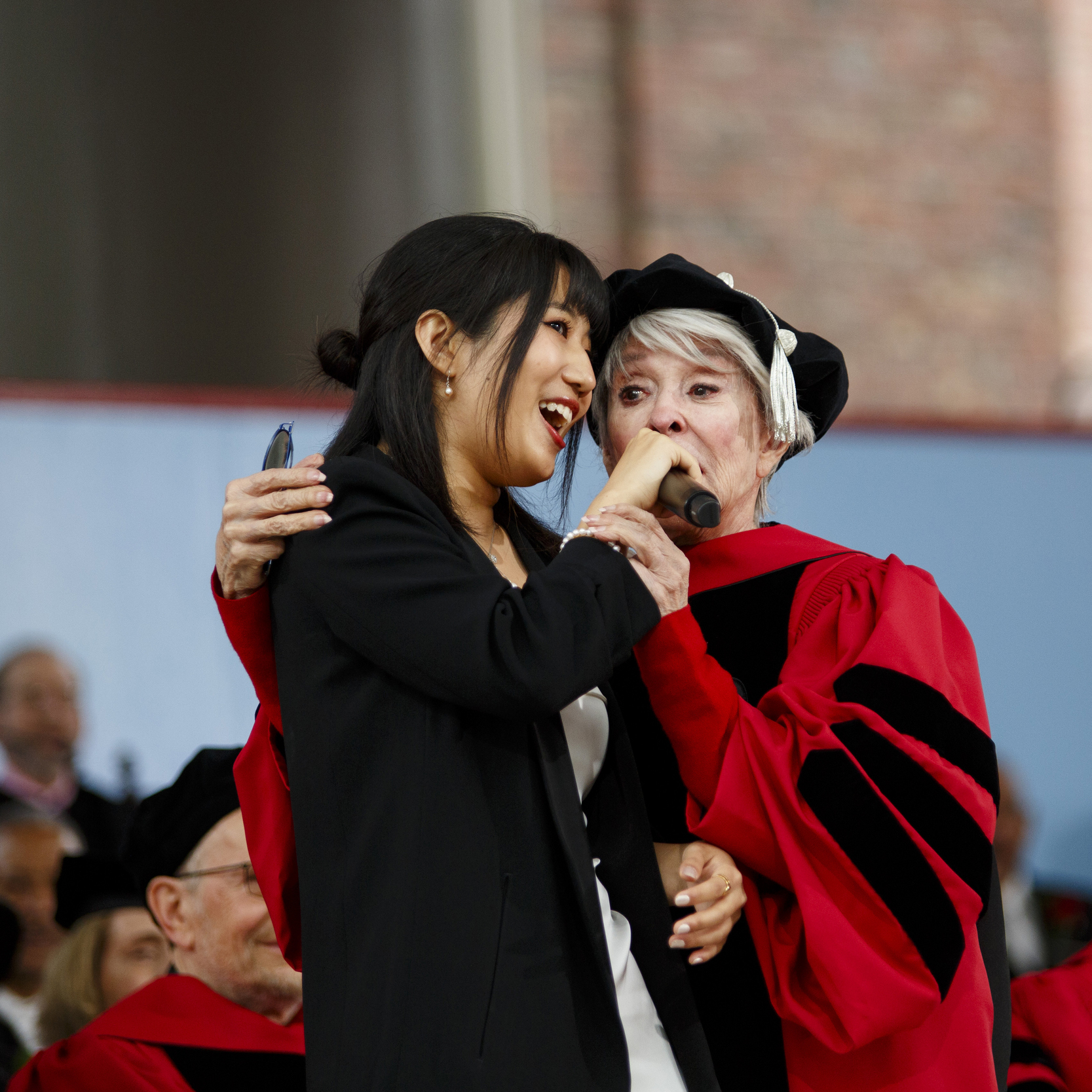 A graduate and an honorary degree recipient sing together on stage.